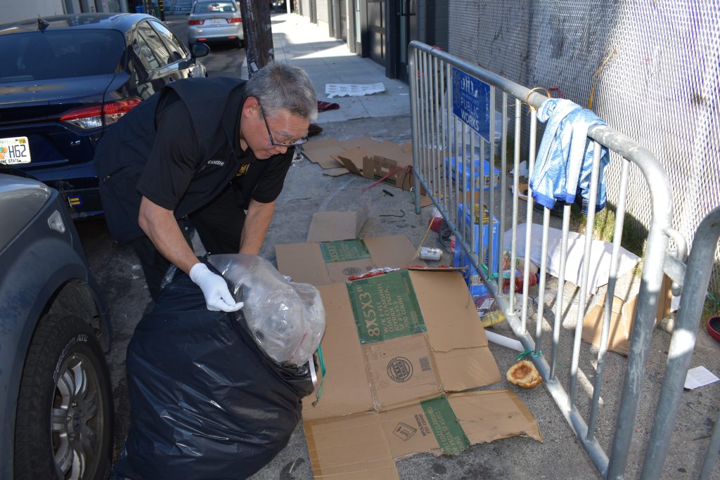 Nakanishi strips apart a solo tent on Stevenson and 14th Street in San Francisco. He discards items, like tarps and cardboard, so that people cannot reuse them to make another sleeping structure. Photo by Magaly Muñoz.