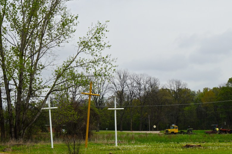 An environmental landscape shows three large metal crosses rising from a lush green field dotted with small white flowers.
