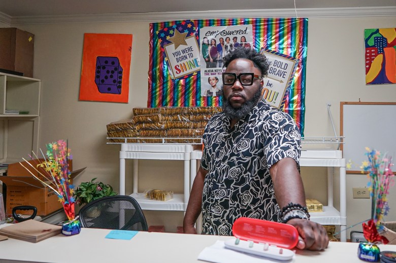 A man standing at the front desk of a small room faces the camera. Behind him are a few signs decorated with rainbows. They say, 
