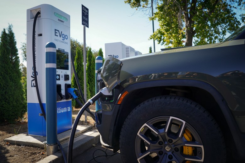 A close-up of an electric vehicle plugged into an EVgo fast charging station in a parking lot. The charging cable connects to the front side of a dark green Rivian truck, with signage for Ultium and EVgo visible in the background.
